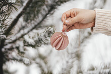 Hand In Cozy Sweater Holding Modern Bauble On Background Of Pine Tree Branches In Snow. Decorating Christmas Tree Outdoors.  Preparation For Winter Holidays In Countryside. Space For Text