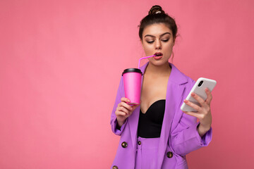 Wall Mural - Shot of concentrated caucasian brunette woman holds take away coffee dressed in casual clothes isolated over pink wall uses smartphone and drinking