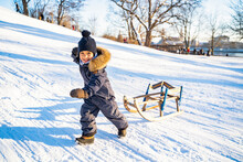 Portrait Of African American Or Latin Joyful Little Boy In Warm Waterproof Overalls, Mittens And Hat Is Pulling Handmade Wooden Sled Along Snow-covered Hill. Sleigh Ride, Holidays, Christmastime