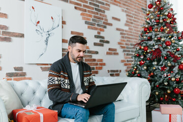 Young man with a beard smiling and working on a laptop from home, sitting on the couch during the New Year holidays