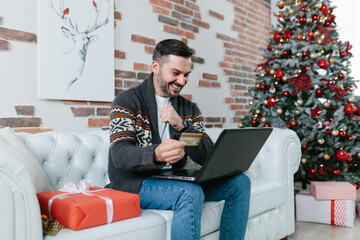 Playful man with a beard smiling sitting on the couch shopping online, buying Christmas presents, online store using laptop