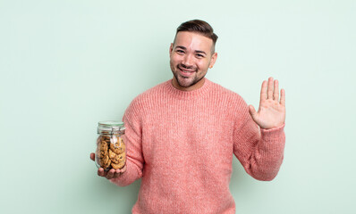 Wall Mural - young handsome man smiling happily, waving hand, welcoming and greeting you. cookies bottle concept