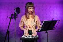 A Young Caucasian Female Learns To Play A Keyboard Instrument On The Notes, Looks Into The Music Stand Next To The Microphone In A Recording Light Studio With Purple Light