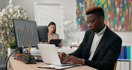 Wall Mural - A smiling dark-skinned boy spends morning at work in front of computer screen, tapping fingers on laptop keyboard, filling out official and company applications, comparing correctness on two monitors