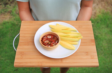 Wall Mural - People hands holding mango slice and sweet fish sauce on white plate on bamboo wooden board.