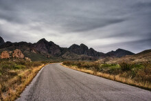 View Of A Road Asphalt Way Between Wild Grass And Black High Rocky Mountains Under A Gray Sky