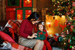 © Elena  - Mother and little daughter open a packed gift box, sitting in the living room decorated for Christmas, celebrating the holiday at home, lights on a blurry background, free space to copy