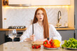 © dikushin - Portrait of attractive redhead young woman with shocked eyes holding big knife sitting at table with cutting board and looking at camera in light kitchen room with modern interior, close-up.