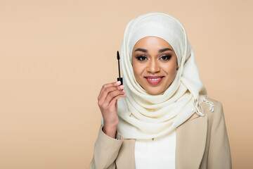 Wall Mural - happy muslim woman in hijab holding mascara brush isolated on beige