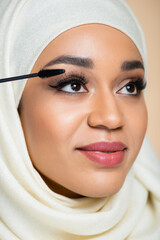 Wall Mural - close up of happy muslim woman in hijab applying mascara with brush isolated on beige