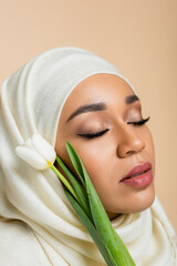 Wall Mural - Portrait of pretty muslim woman in hijab with closed eyes near tulip isolated on beige