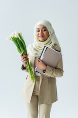 Wall Mural - happy muslim businesswoman in hijab holding tulips and folders isolated on grey