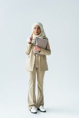 Wall Mural - full length of pensive muslim woman in hijab holding pencil and folders on grey