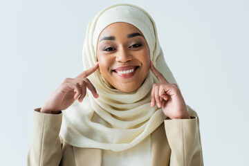 Wall Mural - happy muslim woman in hijab pointing at cheeks while smiling isolated on grey