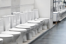 Exhibition Of Samples Of White Toilet Bowls Standing In A Row In The Warehouse Of A Plumbing Store. Samples Of Modern Sanitary Ware For The Toilet. New Modern Toilet In The Plumbing Store
