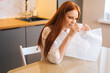 © dikushin - Side view of nervous young woman playing pops bubble wrap to calm herself sitting at table in kitchen. Stressed pretty redhead female popping plastic bubblewrap. Concept of mental health.
