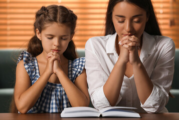 Wall Mural - Young woman with her little daughter praying together over Bible at home