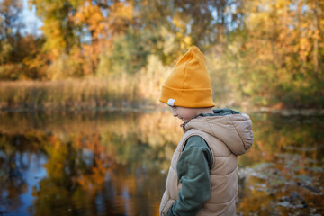 Wall Mural - Pensive little boy stands on the bank of river, admires the magnificent view of autumn forest and its reflection in the lake water. Autumn walk. Outdoor lifestyle, active family lifestyle