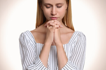 Wall Mural - Religious young woman with clasped hands praying against light background, closeup