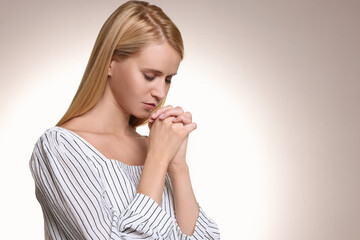 Wall Mural - Religious young woman with clasped hands praying against light background. Space for text