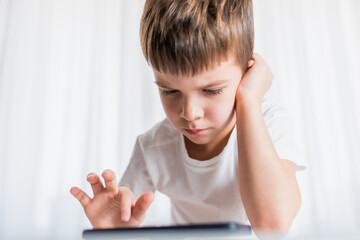 A cheerful child in a white T-shirt plays games on the phone at home. Happy boy looks at his smartphone.