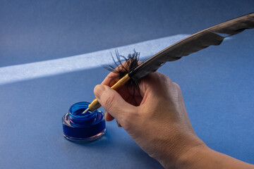 Calligraphic blue ink in a glass inkwell, hand holding a classic black quill pen with a golden metallic nib, deep blue background with beautiful shadows.