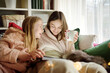 © MNStudio - Two young sisters snuggling up on the sofa in a cozy living room at Christmas. Cute children using a tablet at home during winter break. Kids reading a book in comfy blanket.