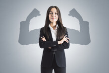 Portrait Of Attractive Young European Businesswoman With Folded Arms And Shadow Muscle Arms On Concrete Wall Background. Strenght And Leadership Concept.