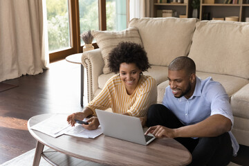 Happy millennial african american family couple analyzing financial paper documents, managing household budget or paying for services in e-banking computer app, enjoying calculating expenses at home.