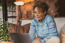 Portrait Of Happy Modern Woman At Home Sitting On The Sofa Using Computer And Wearing Eyewear. Female People Surf The Web And Work With Laptop Smiling. Indoor Technology Leisure Activity Concept