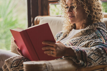 Relaxed adult lady reading a book sitting on the chair at home with outside view. Concept of female people happy lifestyle living alone. Attracvite woman studying indoor