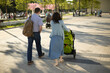 © Олег Копьёв - People walk around the city on a sunny day. Family on a walk. A man with a backpack, in dark trousers and a light shirt. A woman in a skirt and cardigan bluish shades carries a green stroller.