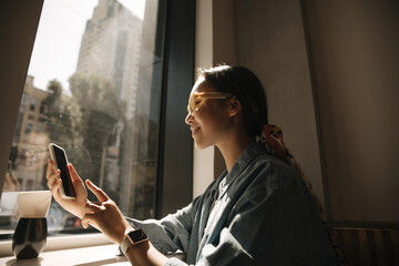 teenage young asian girl using mobile phone, chatting and sending message while sitting near window.