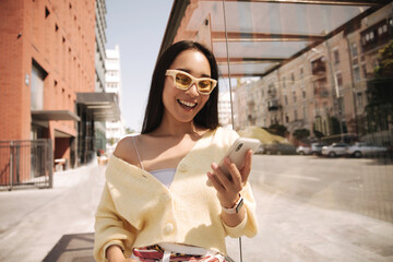 Closeup happy girl looks at phone screen and smiles while standing outdoors. She is thrilled to have received message on her smartphone. Brunette in light casual clothes.