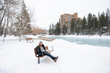 Man Talking On Smart Phone On Snowy Park Bench At Frozen Pond