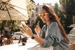 © Look! - Cute young asian brunette in glasses looking at smartphone sitting in cafeteria on street. Happy girl student of university using mobile phone communicates by video communication with friends.