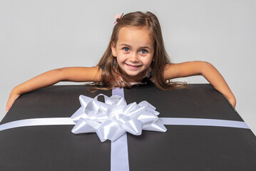 Canvas Print - little cute emotional girl in a colored dress with blond hair leaned on a festive black box with a bow on a white background. She is thoughtful, smiling. Birthday, celebration, anticipation.