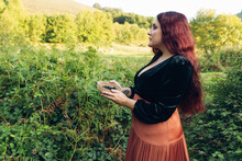 Beautiful Young Woman In A Green Meadow Surrounded By Blackberries And Ferns. She Is Looking To The Side And Holding A Basket Of Ripe Blackberries In Her Hands. Forest Out Of Focus In The Background.