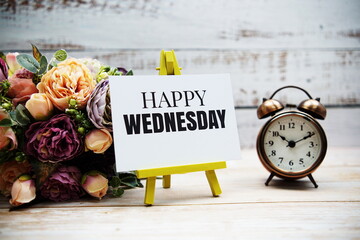 Happy Wednesday text with flower bouquet and alarm clock on wooden background