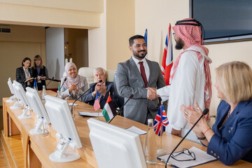 Canvas Print - Smiling mixed race politician in suit making handshake with Arabian representatives in thobe at conference