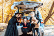 © klavdiyav - siblings boy in skeleton costume, teenage girl in witch costume and hat and cute little girl in spooky costume sits in trunk car decorated for Halloween with web, orange balloons and pumpkins, outdoor