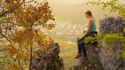 Wall Mural - COPY SPACE: Young female hiker sits on a large rock and observes the lush green valley and emerald river below her. Woman on hiking trip stops to observe the picturesque autumn colored landscape.