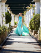 Beautiful blonde milf woman comes in long turquoise flying dress among white columnns along a peristylar outdoor corridor surrounded by green plants and flowers.