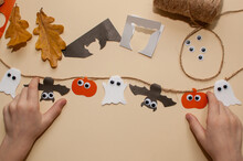 Children's Hands Next To A Garland On A Jute Rope Made Of Pumpkins, Bats And Ghosts With Eyes On A Beige Background With Paper Scraps And Autumn Leaves
