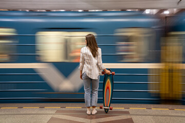 Young girl passenger with longboard standing on subway station platform with blurry moving blue train on background, rear view. Woman with skateboard watching metro pass fast at the station.