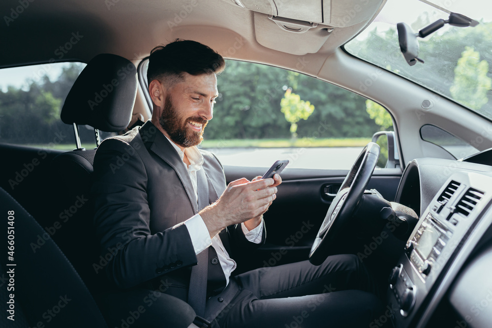 Young man with a beard businessman driving a car in the parking lot smiles and writes on the phone reads the news successful man