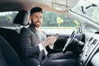 © Liubomir - Young man with a beard businessman driving a car in the parking lot smiles and writes on the phone reads the news successful man