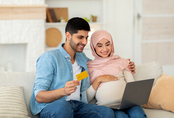 Wall Mural - Preparing for childbirth. Loving muslim pregnant couple using laptop computer and credit card, sitting on sofa