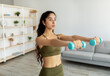 © Prostock-studio - Domestic sports during covid lockdown. Strong Indian woman doing exercises with weights, working out with dumbbells