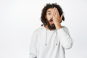 Wall Mural - Portrait of guy covers half of face with palm, looks surprised and amazed at camera with one eye, white background